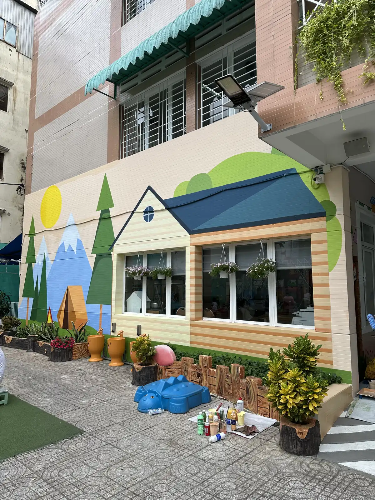 Vẽ tranh tường trường mầm non: mặt tiền ngôi nhà mái xanh, núi và rặng thông